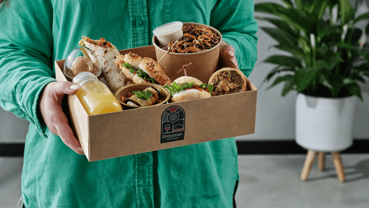 אשה מחזיקה במארז בגודל קופסת נעליים שמכיל מגוון פריטי פינגר פוד סלט ושתיה אישית וקינוח בכוסית