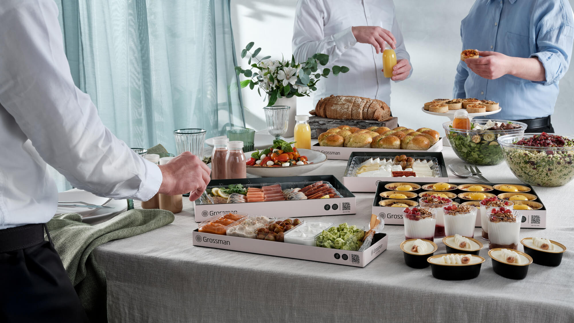 תמונת אווירה של ארוחת בוקר פרימיום, גברים סביב שולחן עמוס במגשי אירוח פרימיום, מתבלים, לחמים, דגים מעושנים וגבינות קשות, סלטים בקערות, עוגות גבינה ומוזלי