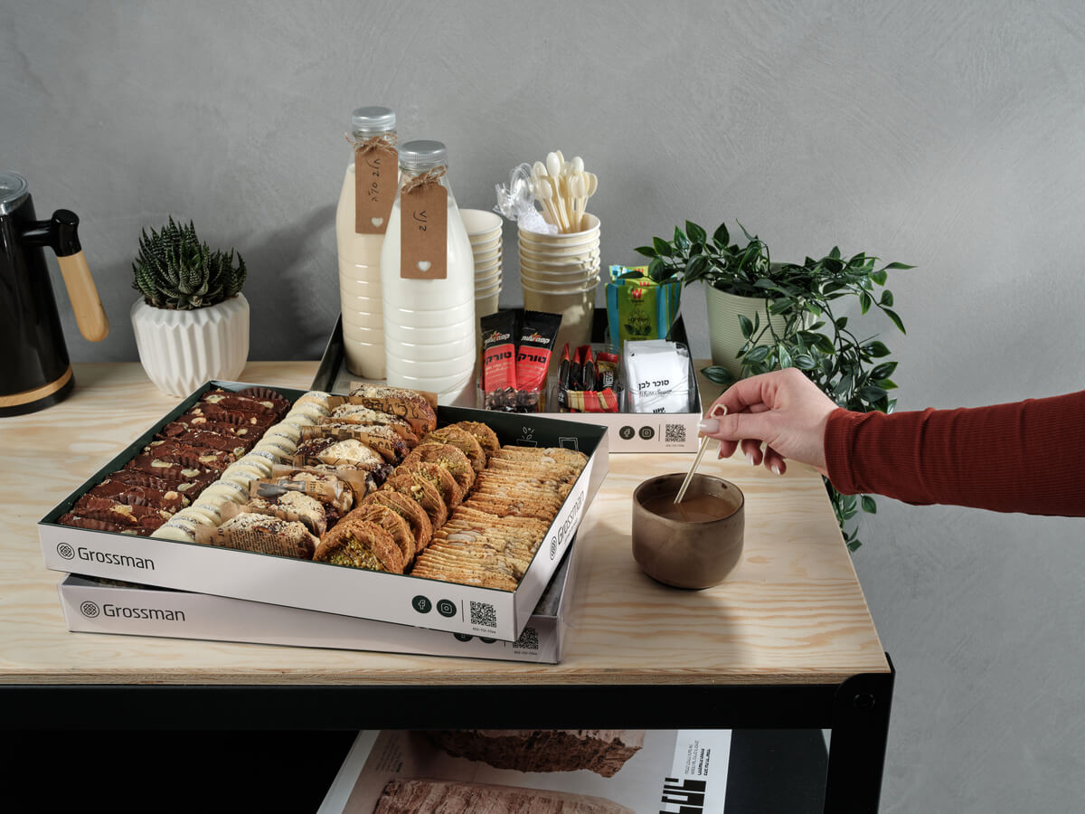 תמונת אווירה של פינת קפה עם מגש אירוח ליד הקפה במגש ישנם 5 סוגים שונים על עוגות ועוגיות. לידו מגש הכנה לקפה ותה.